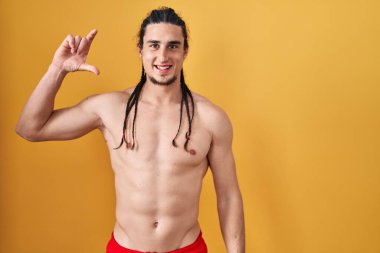 Hispanic man with long hair standing shirtless over yellow background smiling and confident gesturing with hand doing small size sign with fingers looking and the camera. measure concept. 