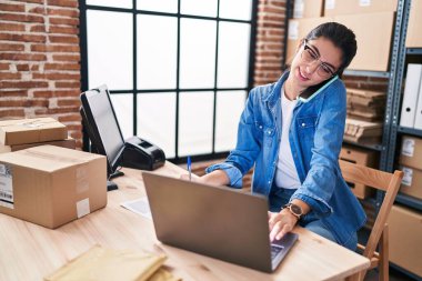 Young beautiful hispanic woman ecommerce business worker using laptop talking on smartphone at office