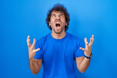 Latin kökenli genç adam mavi arka planda deli deli deli bağırıyor ve agresif bir ifade ve kollarla bağırıyor. hüsran kavramı. 