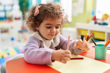 Adorable hispanic girl preschool student sitting on table drawing on paper at kindergarten