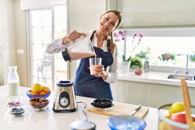 Genç sarışın kadın gülümsüyor. Kendine güvenen. Mutfaktaki cama meyve suyu döküyor.