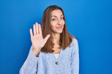 Mavi arka planda duran genç bayan merhaba diyor mutlu, güler yüzlü, dostça karşılama jesti 