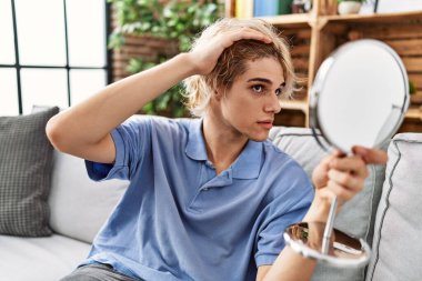 Young blond man looking hair on mirror sitting on sofa at home