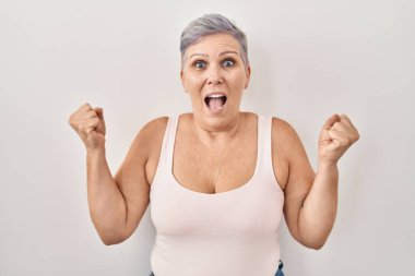 Beyaz arka planda duran orta yaşlı beyaz kadın kollarını kaldırıp, gözlerini açarak başarının şaşkınlığını ve şaşkınlığını kutluyor. Kazanan konsept. 