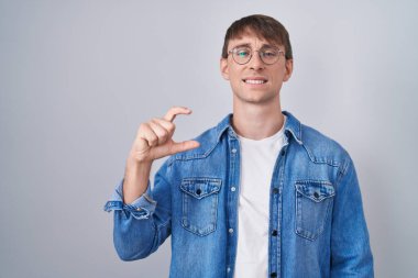 Caucasian blond man standing wearing glasses smiling and confident gesturing with hand doing small size sign with fingers looking and the camera. measure concept. 