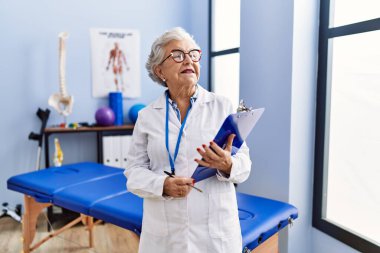 Fizyoterapi kliniğinde fizyoterapist üniforması giyen yaşlı, gri saçlı bir kadın kontrol listesi tutuyor.