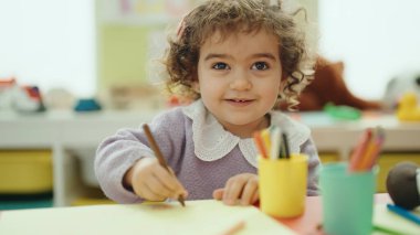 Adorable hispanic girl preschool student sitting on table drawing on paper at kindergarten