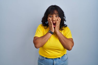 Mavi arka plan üzerinde duran İspanyol kadın korkmuş ve şok olmuş, şaşırmış ve şaşırmış bir yüz ifadesi var. 