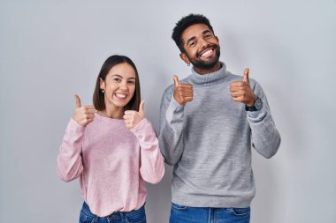 Young hispanic couple standing together success sign doing positive gesture with hand, thumbs up smiling and happy. cheerful expression and winner gesture. 