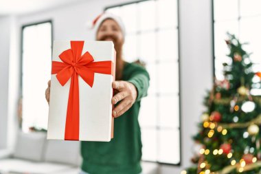 Genç kızıl saçlı adam elinde hediyeyle Noel ağacının yanında duruyor.