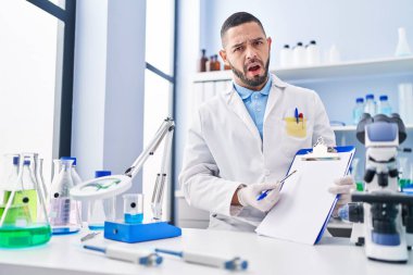 Hispanic man working at scientist laboratory holding blank clipboard in shock face, looking skeptical and sarcastic, surprised with open mouth 