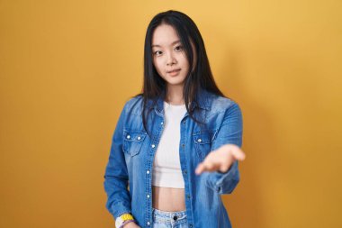 Young asian woman standing over yellow background smiling cheerful offering palm hand giving assistance and acceptance. 