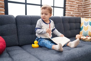 Sevimli beyaz çocuk kanepede oturmuş defterine çizim yapıyor.