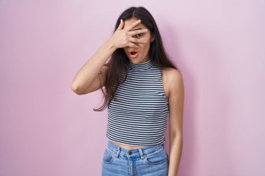 Young teenager girl wearing casual striped t shirt peeking in shock covering face and eyes with hand, looking through fingers with embarrassed expression. 