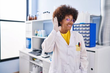 Young african american woman working at scientist laboratory smiling with hand over ear listening and hearing to rumor or gossip. deafness concept. 
