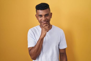 Sarı arka planda, elleri çenesinde soru sormayı düşünen genç İspanyol bir adam. Düşünceli bir ifade. Gülümseyen ve düşünceli bir yüz. şüphe kavramı. 