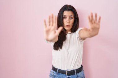 Genç esmer kadın pembe arka planda duruyor. Avuçlarıyla jestleri durduruyor. Öfkeli ve hayal kırıklığı dolu bir ifade. 