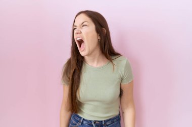 Beautiful brunette woman standing over pink background angry and mad screaming frustrated and furious, shouting with anger. rage and aggressive concept. 
