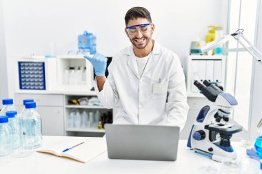 Laboratuvarda çalışan genç İspanyol bir adam dizüstü bilgisayarla parmak ucunda ağzı açık bir şekilde gülümsüyor. 