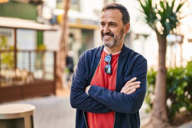 Orta yaşlı bir adam gülümsüyor. Kollarını kavuşturup caddede duruyor.