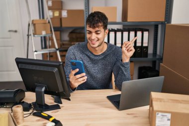 Young hispanic man working at small business ecommerce using smartphone smiling and looking at the camera pointing with two hands and fingers to the side. 