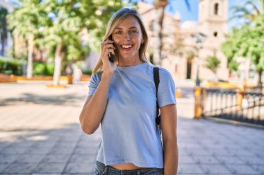 Genç sarışın kadın gülümsüyor. Parktaki akıllı telefondan konuşuyor.