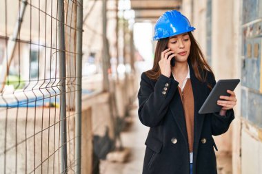Young beautiful hispanic woman architect using touchpad talking on smartphone at street
