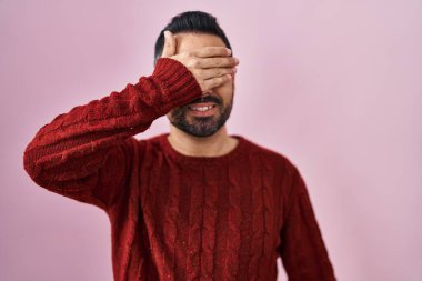 Sakallı, pembe arka planda sıradan bir süveter giyen, gülümseyen ve gülen genç İspanyol bir adam şaşırmak için gözlerini kapatıyor. kör kavram. 