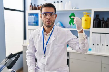 Young hispanic man with beard working at scientist laboratory holding green ribbon scared and amazed with open mouth for surprise, disbelief face 