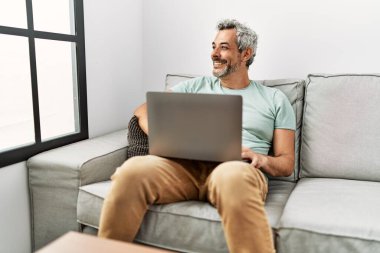 Evdeki koltukta oturan orta yaşlı, kır saçlı adam dizüstü bilgisayar kullanıyor.