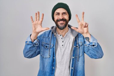 Young hispanic man with tattoos wearing wool cap showing and pointing up with fingers number eight while smiling confident and happy. 