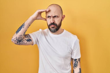 Sarı arka planda dikilen İspanyol dövmeli genç adam. Alnındaki bir sorun yüzünden endişeli ve stresli. Kriz için endişeli. 