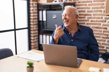 Senior man with grey hair working using computer laptop at the office smiling with happy face looking and pointing to the side with thumb up. 