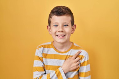 Yüzünde mutlu ve doğal bir ifadeyle el ve parmak ile işaret eden bir gülümseme ile sarı arka planda duran beyaz çocuk. 