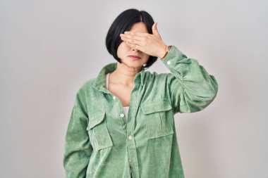 Young asian woman standing over white background covering eyes with hand, looking serious and sad. sightless, hiding and rejection concept 
