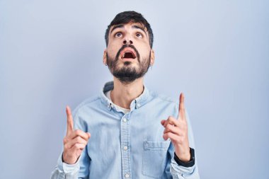 Mavi arka planda dikilen sakallı genç İspanyol adam şaşırmış ve şaşırmış bir şekilde yukarı baktı ve parmaklarıyla işaret ederek kollarını kaldırdı.. 