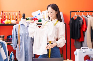 Young chinese woman tailor smiling confident holding t shirt at atelier