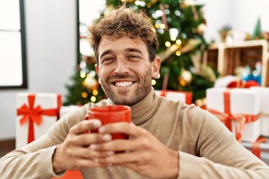 Genç İspanyol adam evde Noel ağacının yanında kahve içiyor.