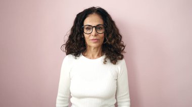 Middle age hispanic woman standing with serious expression over isolated pink background