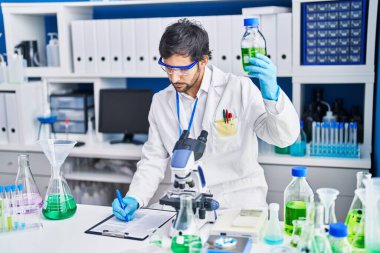 Young hispanic man scientist write on clipboard holding bottle at laboratory