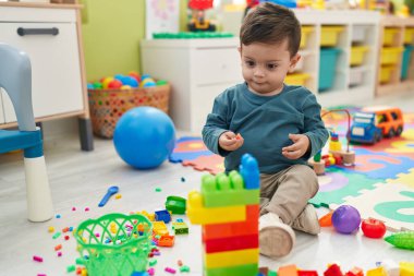 Adorable hispanic boy playing with construction blocks sitting on floor at kindergarten