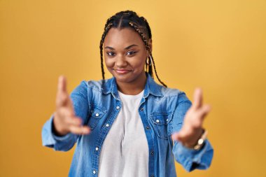 African american woman with braids standing over yellow background smiling cheerful offering hands giving assistance and acceptance. 