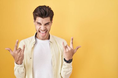 Young hispanic man standing over yellow background shouting with crazy expression doing rock symbol with hands up. music star. heavy music concept. 