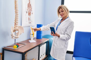 Middle age blonde woman wearing physiotherapist uniform using smartphone pointing to anatomical model of knee at physiotherapy clinic