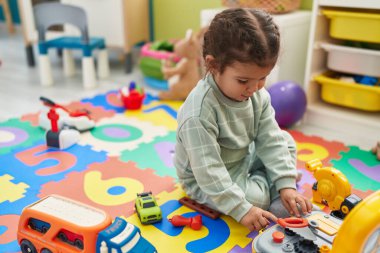 Adorable hispanic toddler playing with car toy sitting on floor at kindergarten