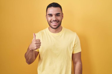 Sarı arka planda dikilen İspanyol genç adam elleriyle mutlu bir jest yapıyor. Kameraya bakıp başarıyı gösteren ifade. 