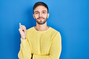 Hispanic man standing over blue background pointing with hand finger to the side showing advertisement, serious and calm face 