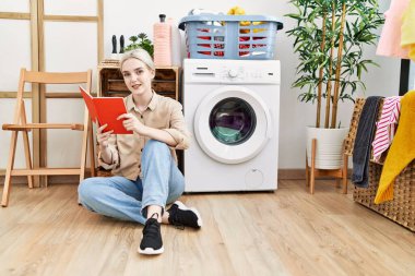 Çamaşır odasında çamaşır makinesi bekleyen, kitap okuyan beyaz bir kadın.