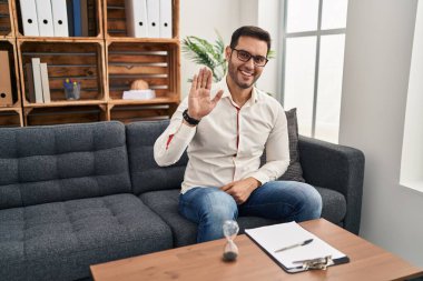 Young hispanic man with beard working at consultation office waiving saying hello happy and smiling, friendly welcome gesture 