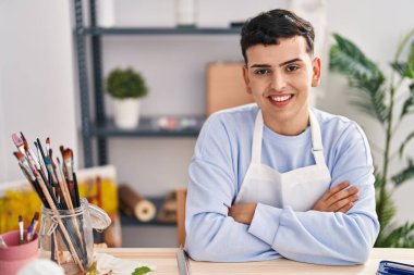 Young non binary man artist smiling confident sitting with arms crossed gesture at art studio